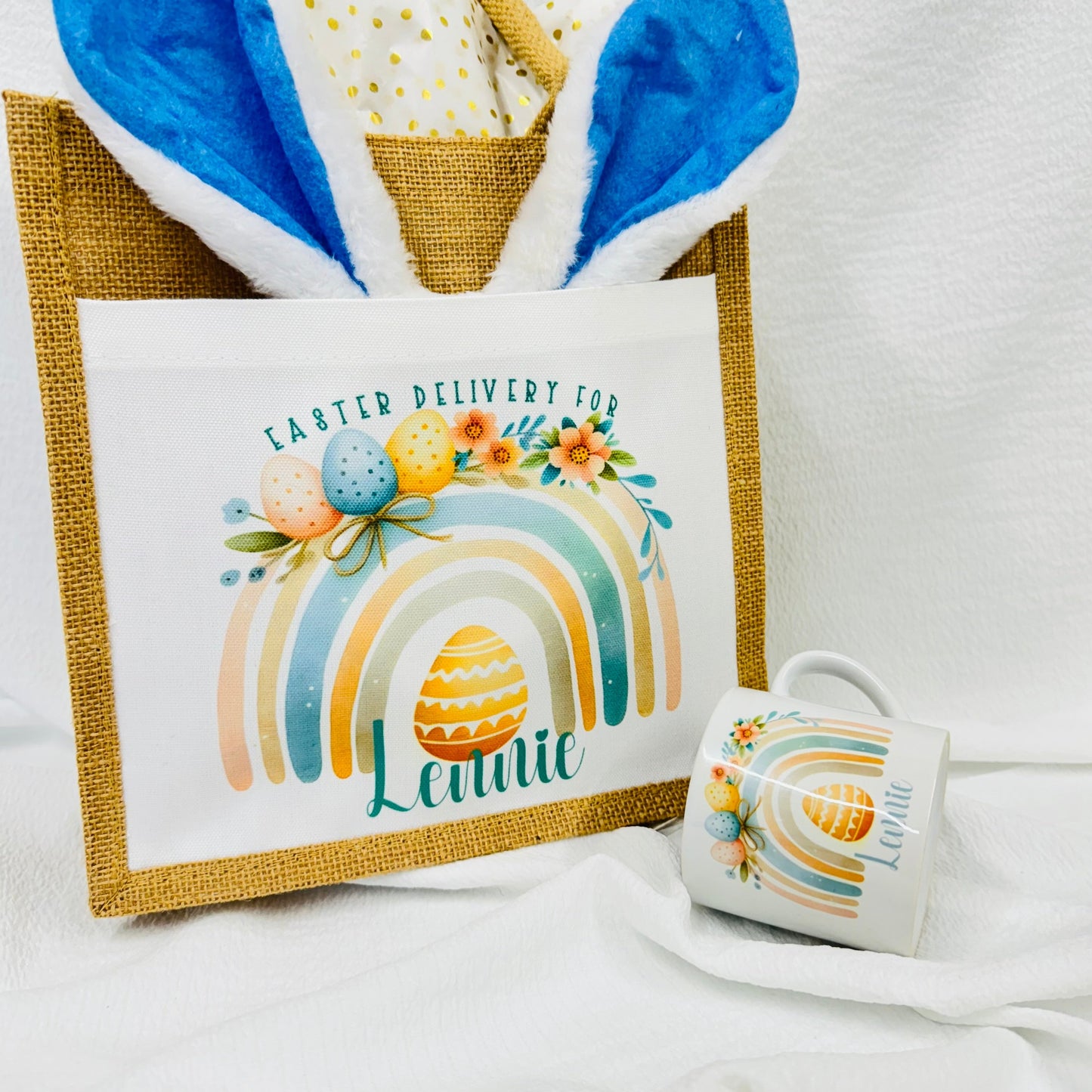 This is a jute gift bag. Printed on the front of the bag is a pastel rainbow decorated with easter eggs and leaves. A name is printed at the bottom of the rainbow. The name can be printed in pink or blue.