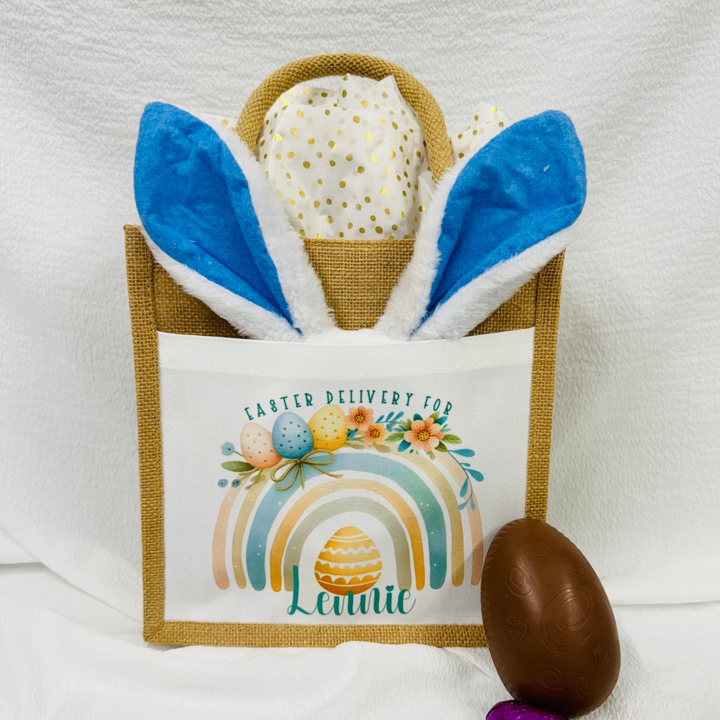This is a jute gift bag. Printed on the front of the bag is a pastel rainbow decorated with easter eggs and leaves. A name is printed at the bottom of the rainbow. The name can be printed in pink or blue.