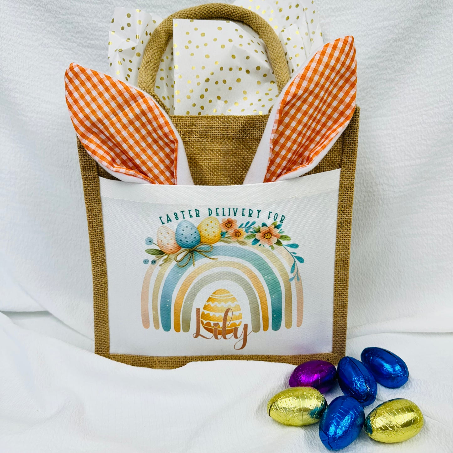 This is a jute gift bag. Printed on the front of the bag is a pastel rainbow decorated with easter eggs and leaves. A name is printed at the bottom of the rainbow. The name can be printed in pink or blue.