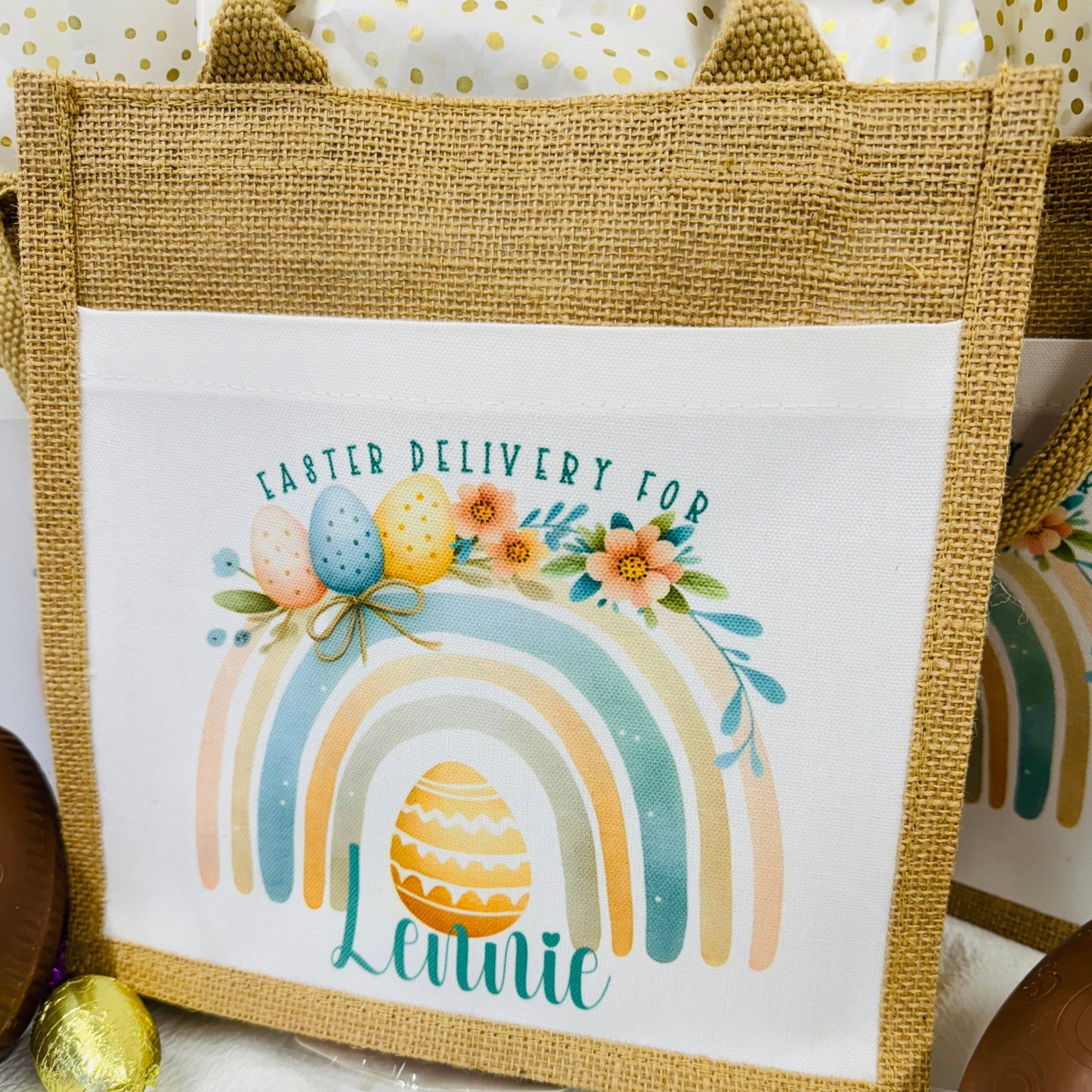 This is a jute gift bag. Printed on the front of the bag is a pastel rainbow decorated with easter eggs and leaves. A name is printed at the bottom of the rainbow. The name can be printed in pink or blue.