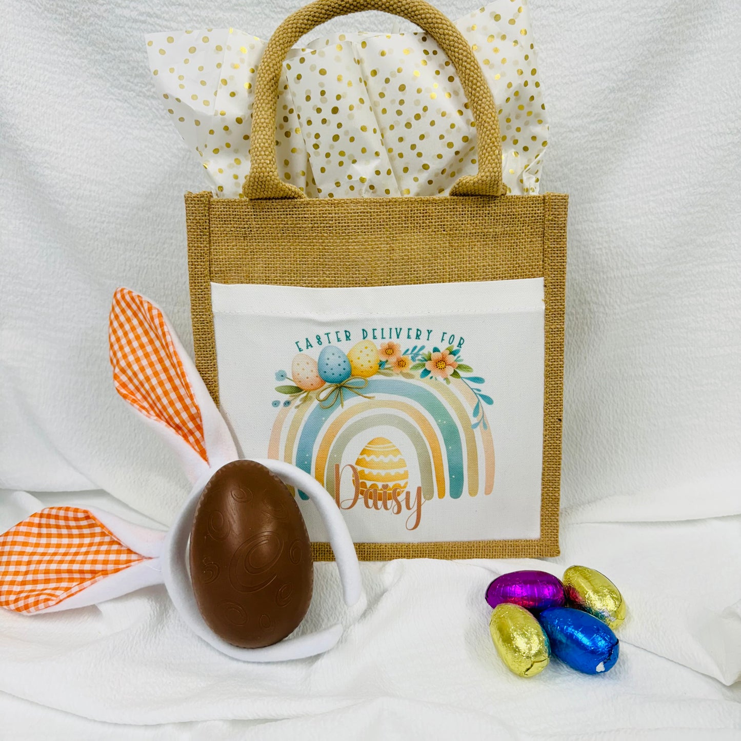 This is a jute gift bag. Printed on the front of the bag is a pastel rainbow decorated with easter eggs and leaves. A name is printed at the bottom of the rainbow. The name can be printed in pink or blue.