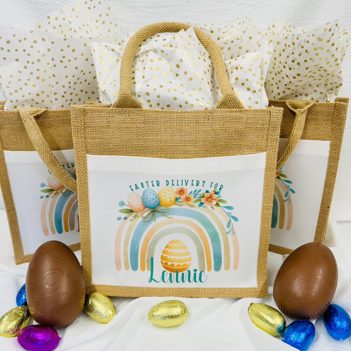 This is a jute gift bag. Printed on the front of the bag is a pastel rainbow decorated with easter eggs and leaves. A name is printed at the bottom of the rainbow. The name can be printed in pink or blue.