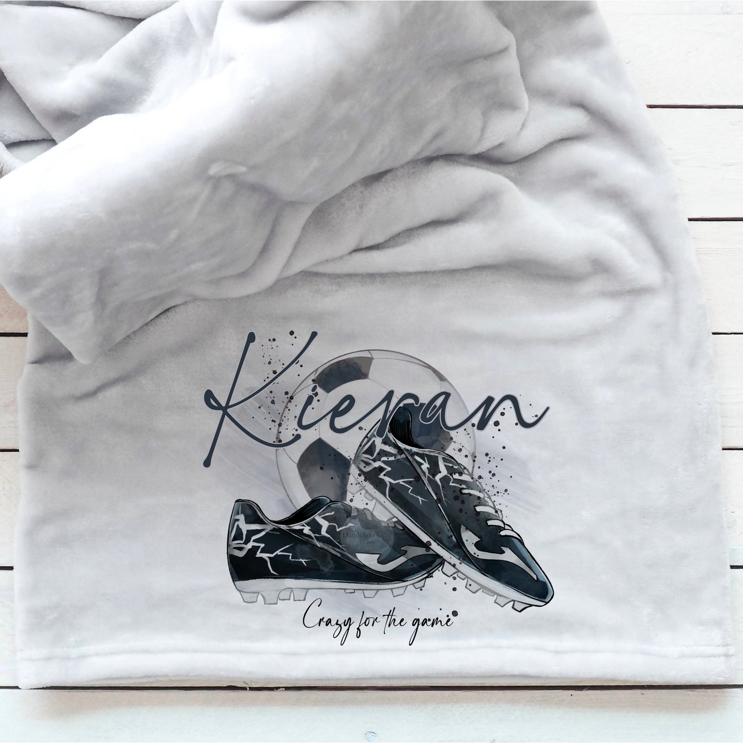 This is a luxury, thick grey fleece blanket. The bottom of the blanket has been printed with a watercolour design of a football with boots resting against it. A name is printed across the top of the whole image. Football fan gift idea.