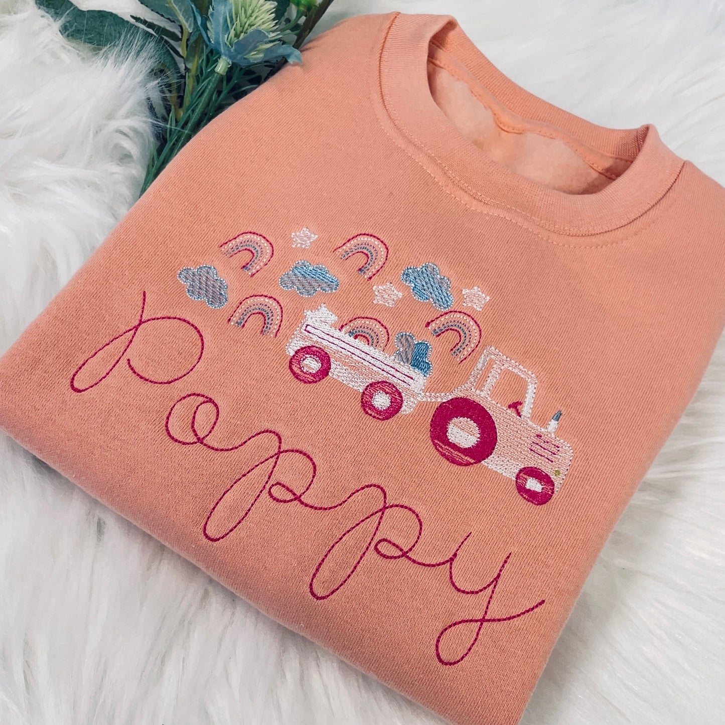 This is a cute pink sweater for girls. It is personalised on the front with an embroidered tractor and trailer carrying rainbows and clouds. A name is also embroidered below the main design. The sweater is cotton with a fleece lining.