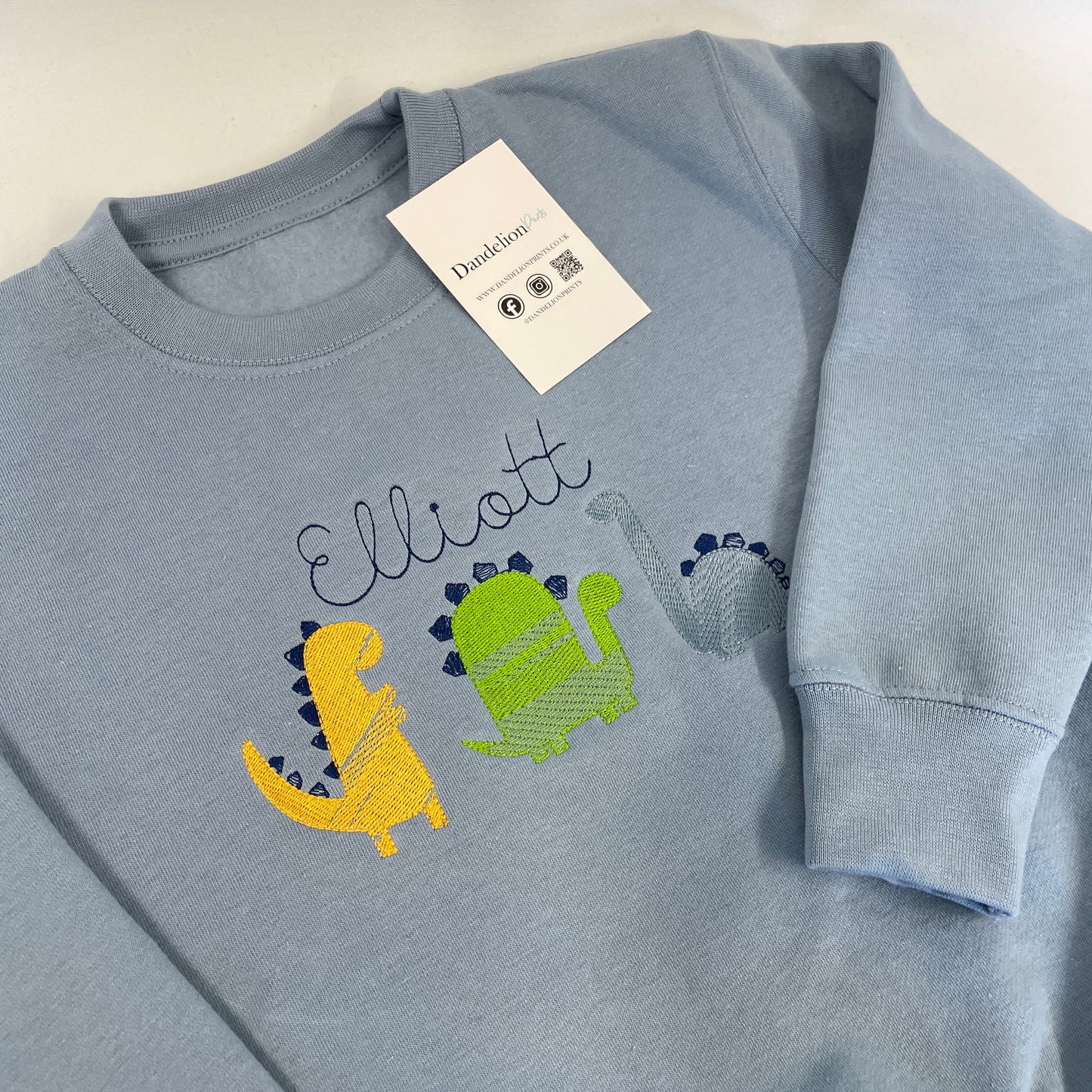 This is a blue sweater with a trio of dinosaurs embroidered on the front. The dinosaurs are embroidered in yellow, green and blue in a sketch style stitch. There is also a name embroidered above the dinosaurs