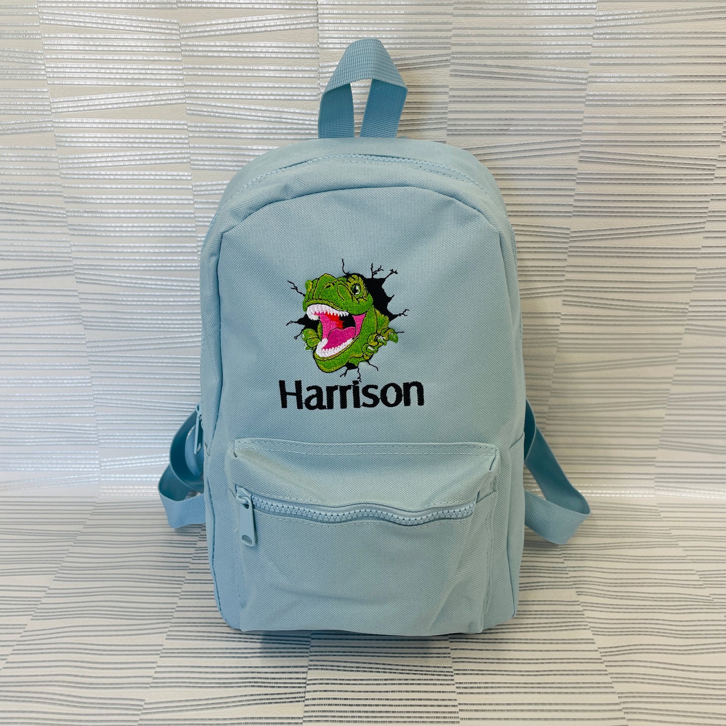 This is a sky blue backpack with a T-Rex embroidered on the front. The T-Rex looks as lough it is bursting out of the bag. There is a name below the embroidery. The bags are available in navy, sage, pink, lilac, grey, mint and sky bljue.