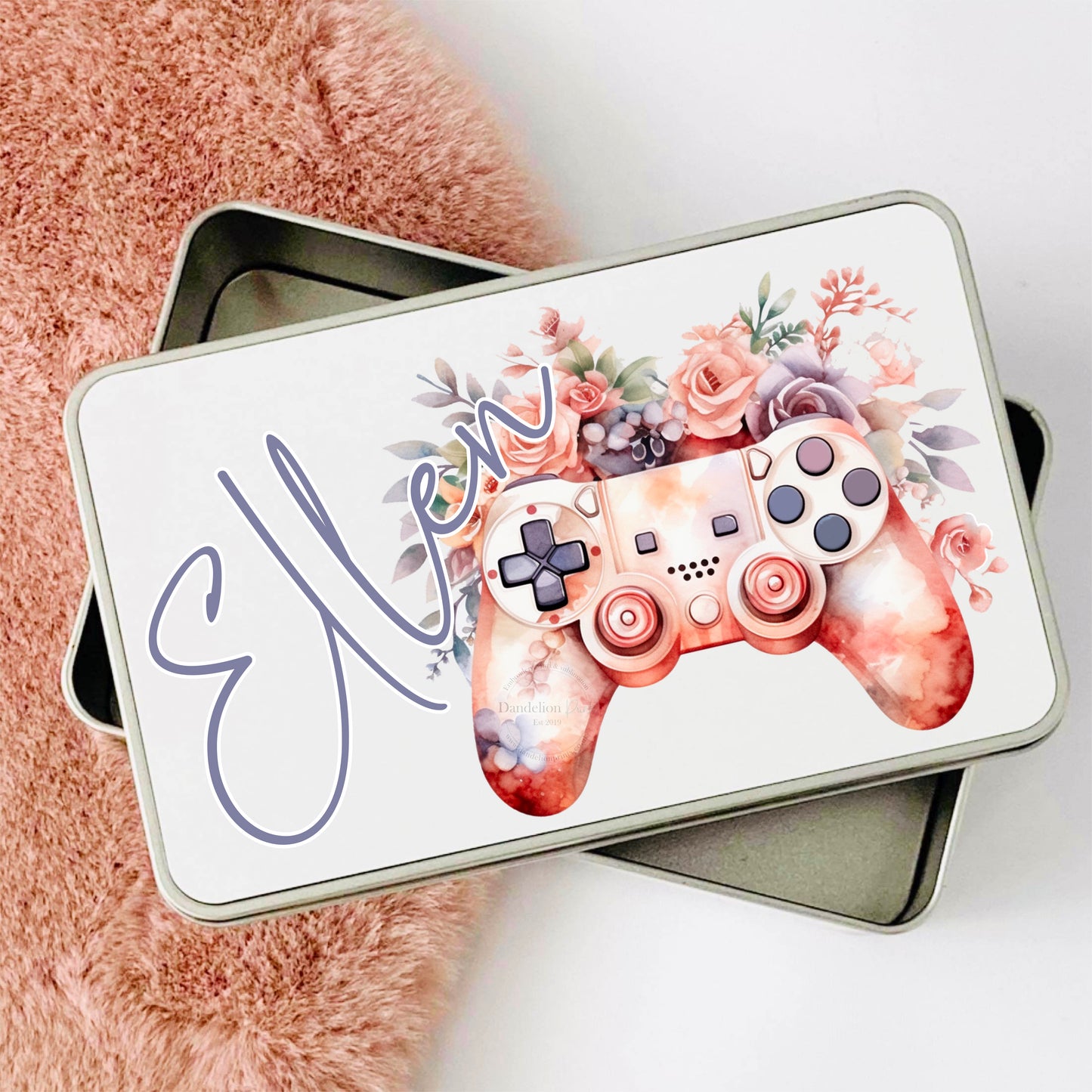 This is a metal storage or accessory tin. Perfect for storing little gadgets and niknaks. On the lid of the tin is a pink floral gaming controller. the flowers are pink and purple. A name is also printed in purple across the tin.