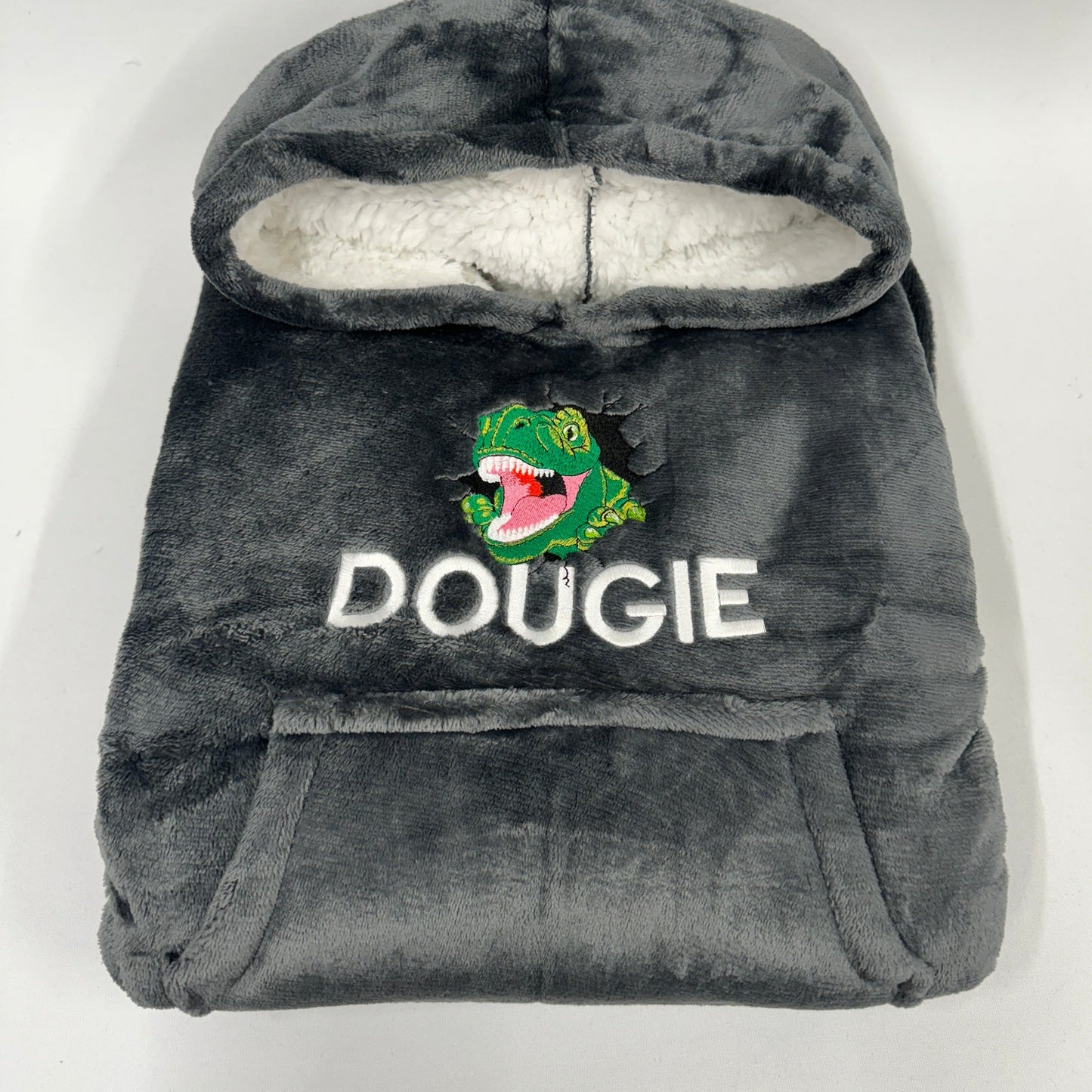 This is a sherpa oversized oodie style hoodie. On the front of the hoodie is an embroidered T-Rex bursting out of the hoodie. There is also a name embroidered below the dinosaur. This is a great gift for any dinosaur lover adult and child!