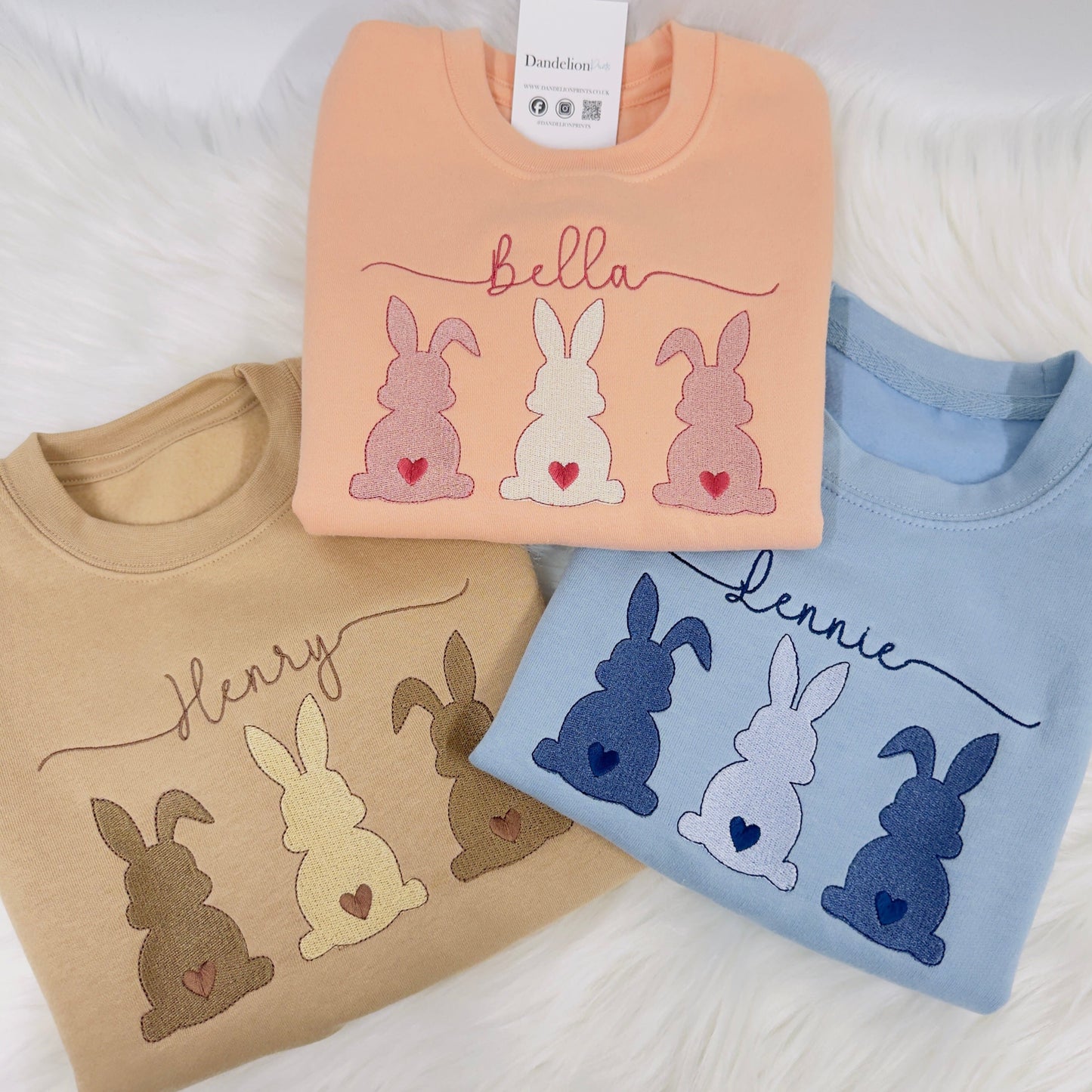 In this photo is three children’s sweaters in blue, pink and taupe. The sweaters have three bunnies in a row. The bunnies are the same colours as the sweaters but different shades. A decorative name is at the top. Easter bunny ready!