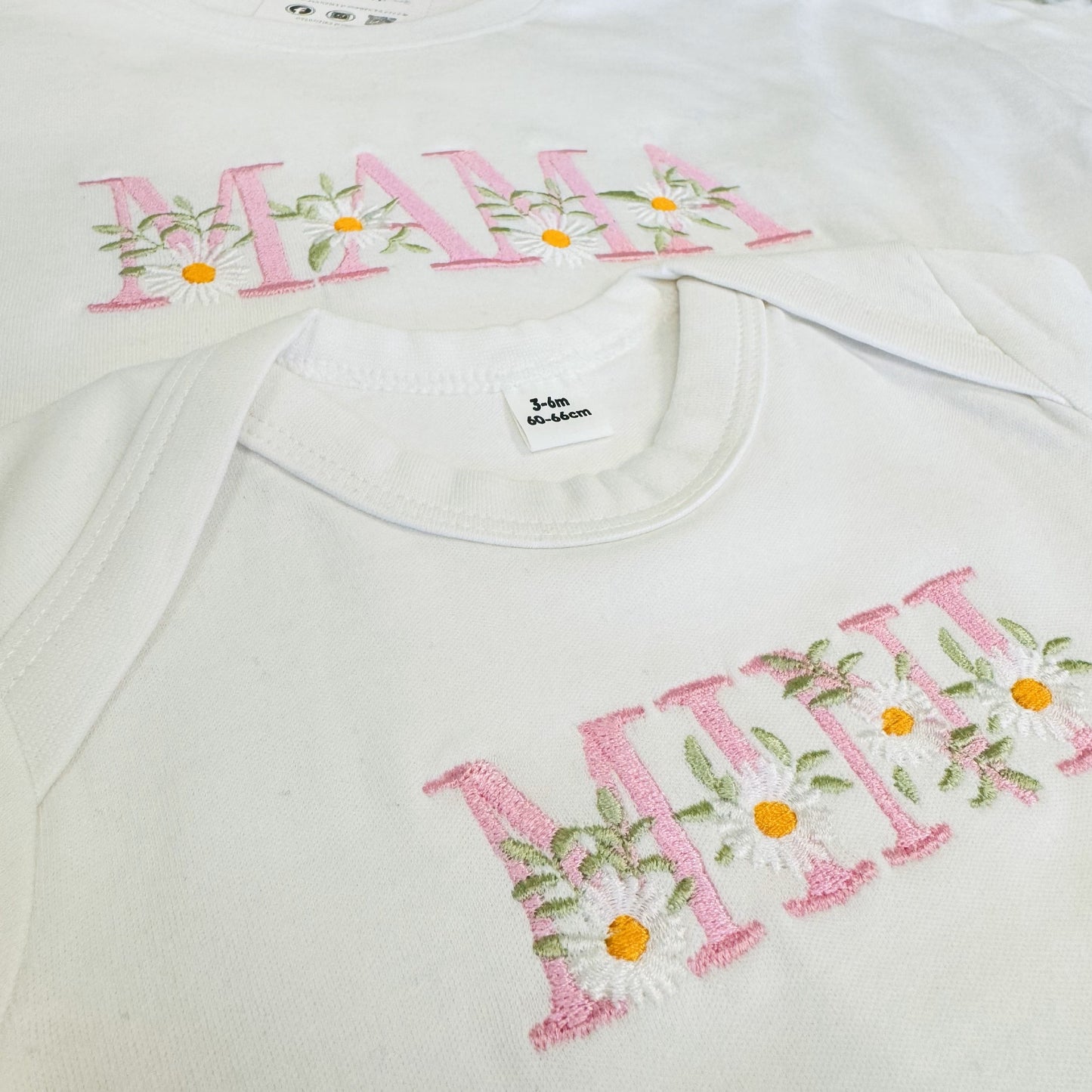 This is a photo of two matching t-shirts. One t-shirt is for an adult and has embroidered on the chest MAMA with flowers. The other t-shirt is for a baby which has MINI embroidered with flowers too. Matching set for mum and baby