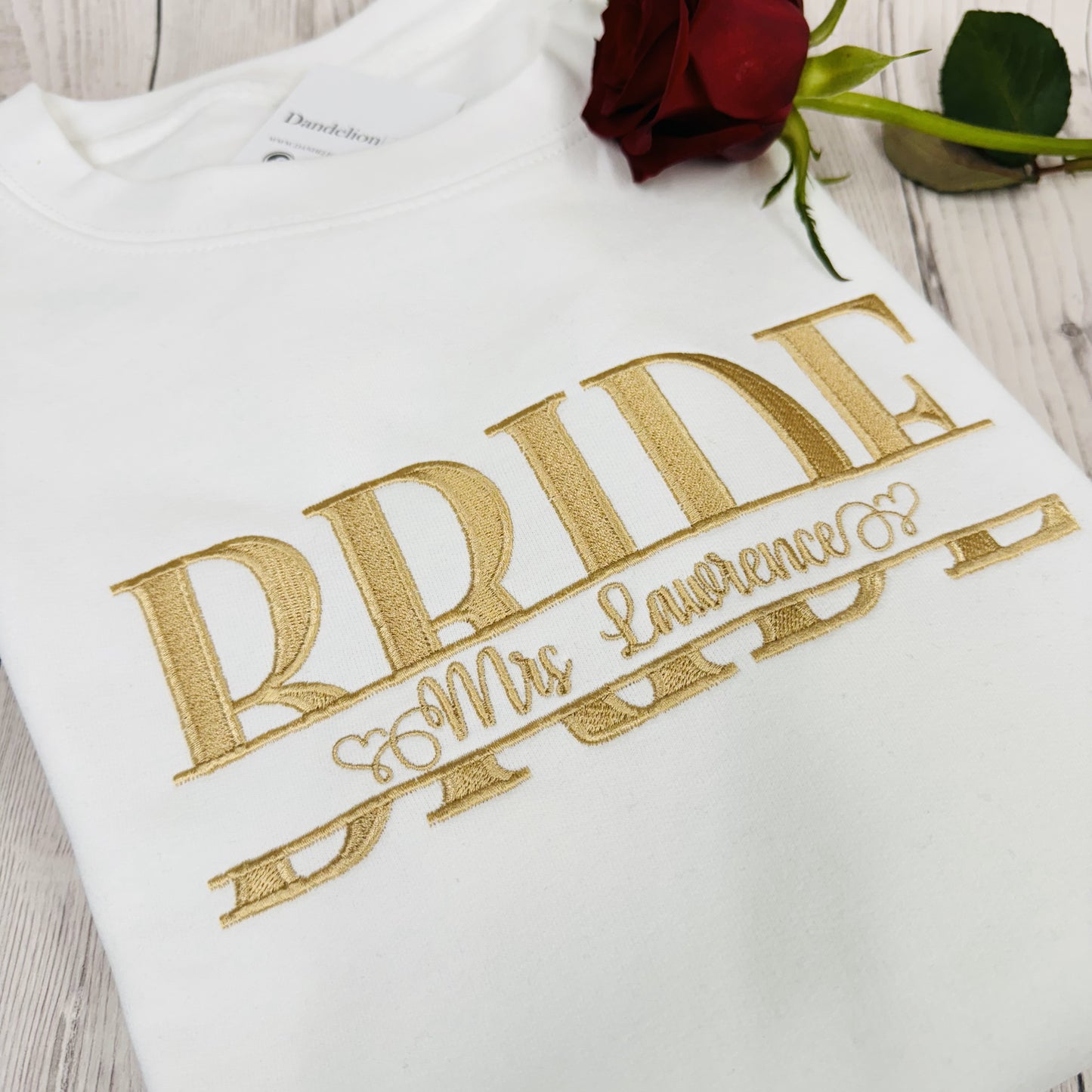 This is a photo of a white sweatshirt with BRIDE embroidered across the chest. The BRIDE is a monogram with a name inside. The embroidery is delicately stitched in an elegant gold thread.