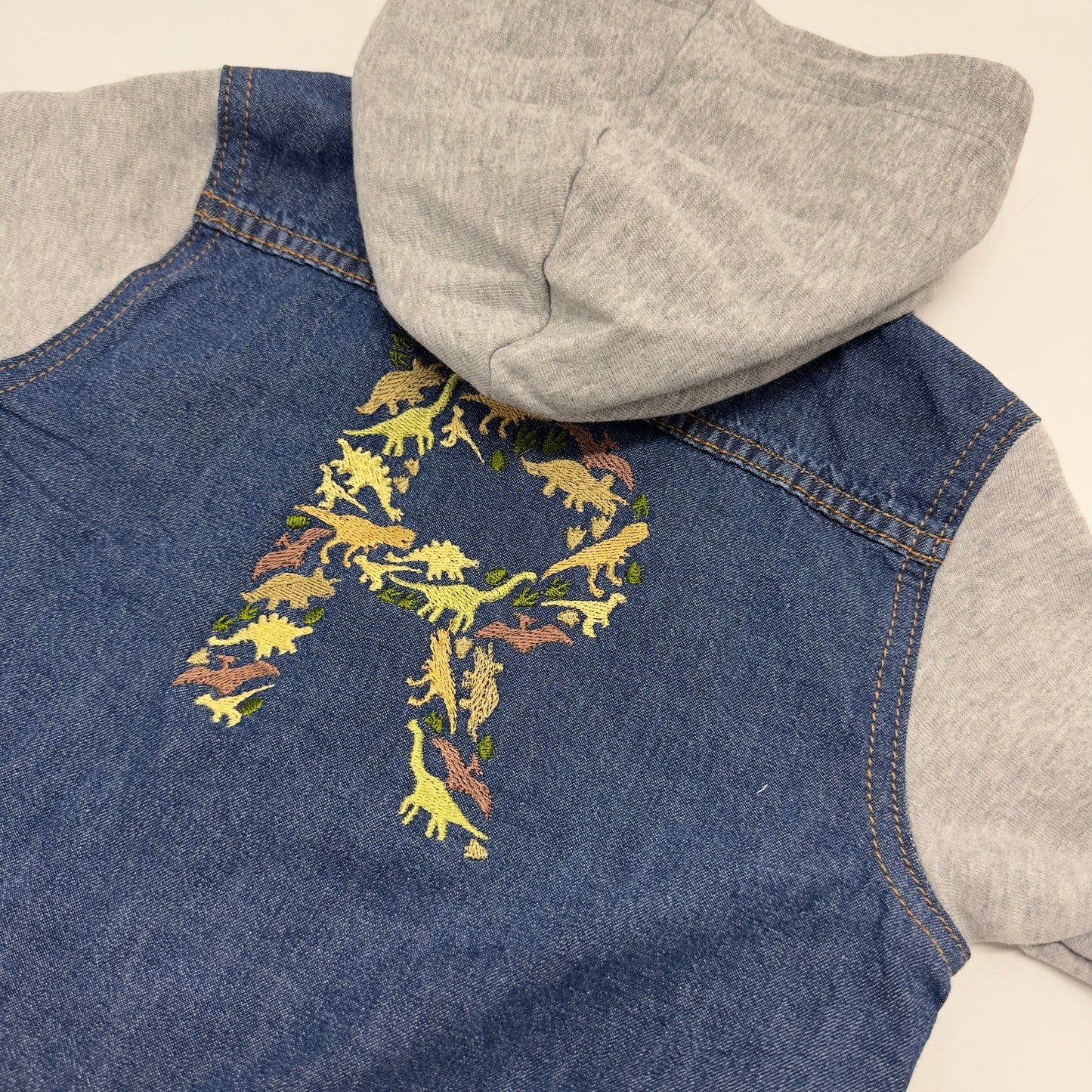 Introducing our trendy Dino Jacket with Cotton Sleeves and Hood!
If you would like your embroidery in a different colour to the greens shown please add this to your personalisation.
This versatile piece combines the classic 100% cotton denim style with cosy cotton sleeves and a hood for added comfort.
The two front pockets allow for convenient storage, while the popper closing ensures a secure fit.
The highlight of this jacket is the embroidered dinosaur initial on the back, adding a whimsical touch to its