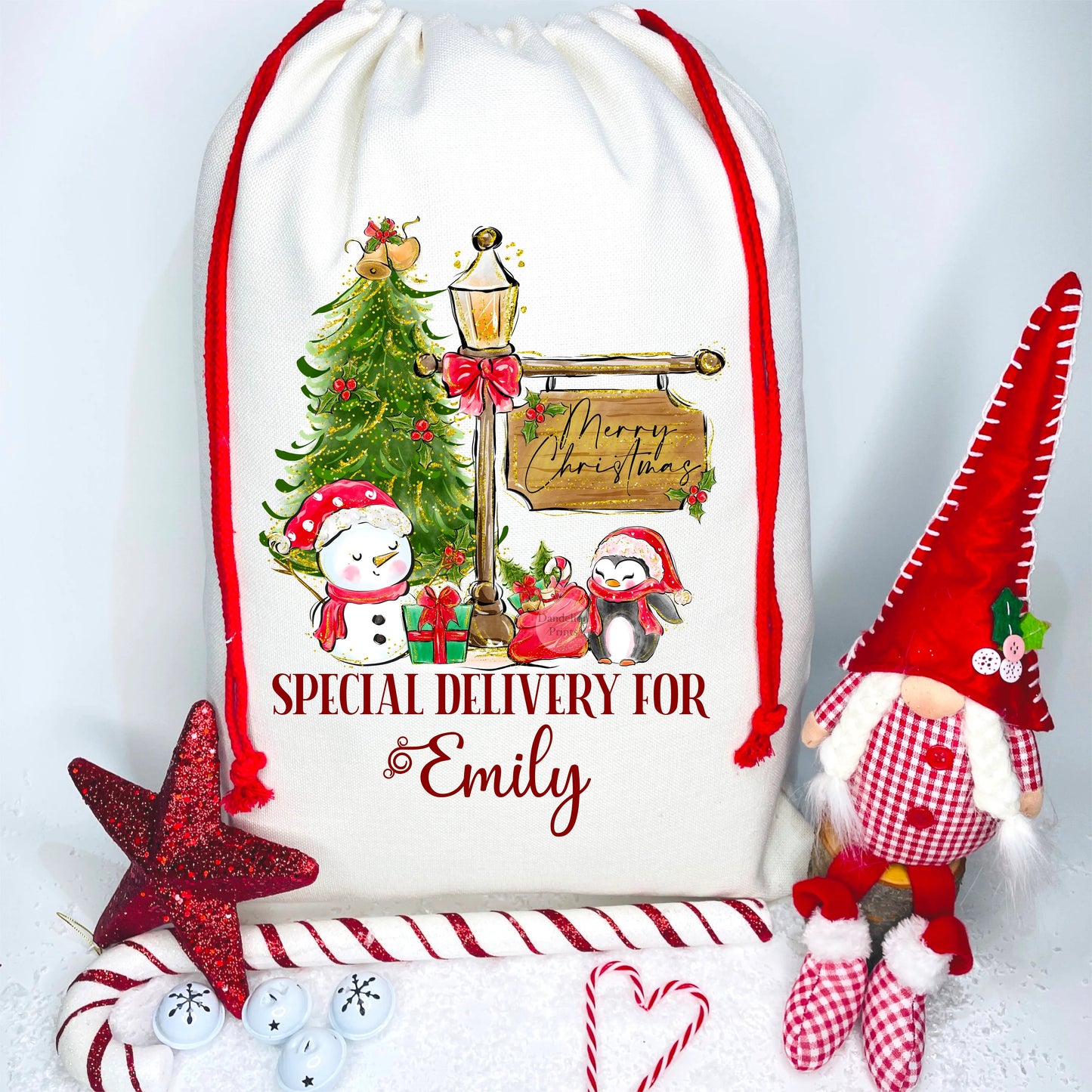 This is a Christmas sack printed with a snowman and penguin wearing hats and scarves, beside a Christmas tree and lamppost with a hanging sign. Featuring presents and a Santa sack. At the bottom is writing says Special Delivery For, with a name.