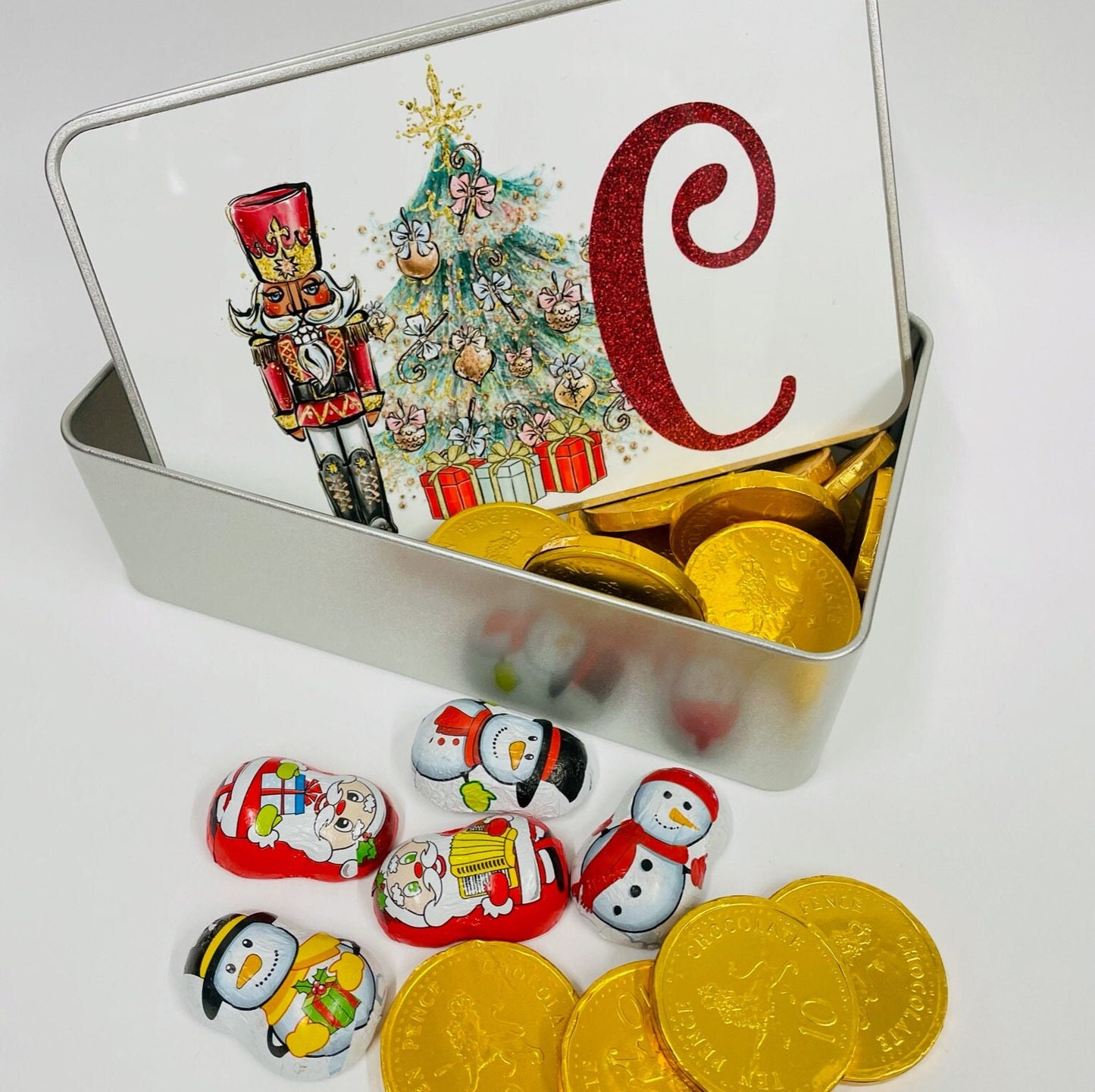 This is a white topped tin printed with a red nutcracker standing beside a Christmas tree which is decorated with pastel colours and presents under the tree. Next to the tree is a large glittery red initial.