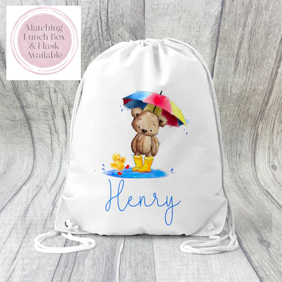 Image of a white PE bag with white drawstrings and a printed image to the front. The image is of a teddy bear wearing wellies, holding an umbrella and stood in a puddle next to a small yellow duck. below is the name Henry.