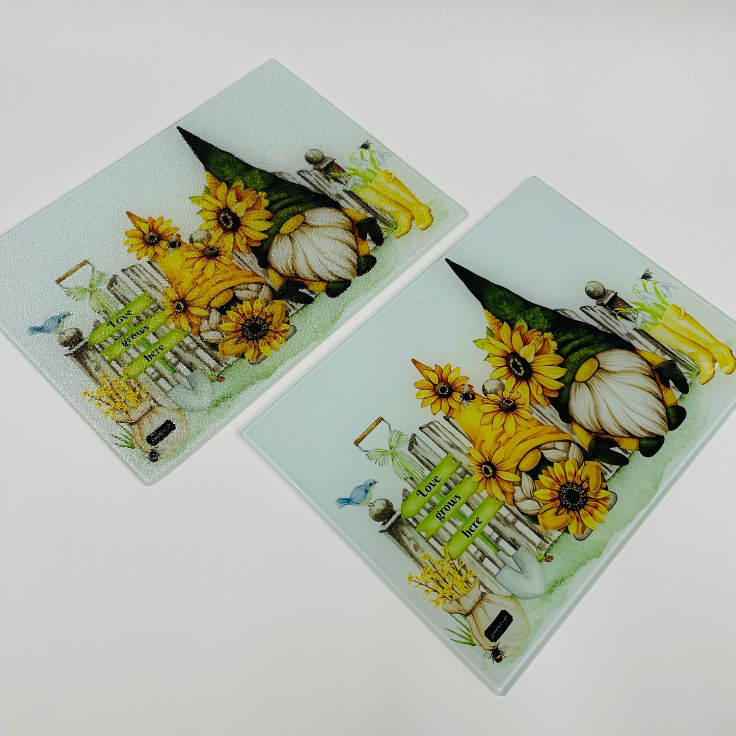 This is a glass chopping board in A4 or A3 size. The design is printed on the reverse which features two gonks with sunflower hats surrounded by garden accessories. With a garden sign that reads Love grows here.