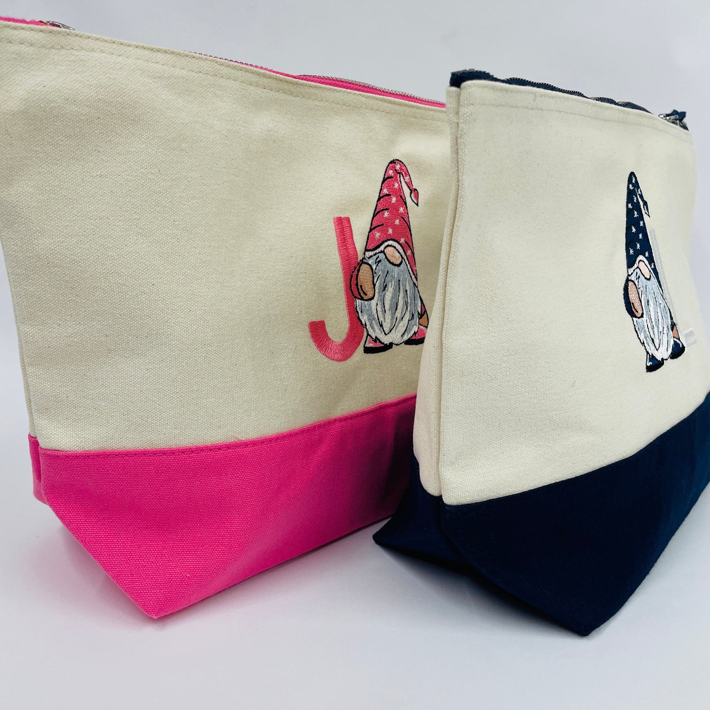 Two personalised make up accessory bags. The main colour of the bags is linen cream, one has a pink zip and pink base. The other has a navy zip and navy base. Each bag is embroidered with an initial and a pink or blue gonk/gnome.