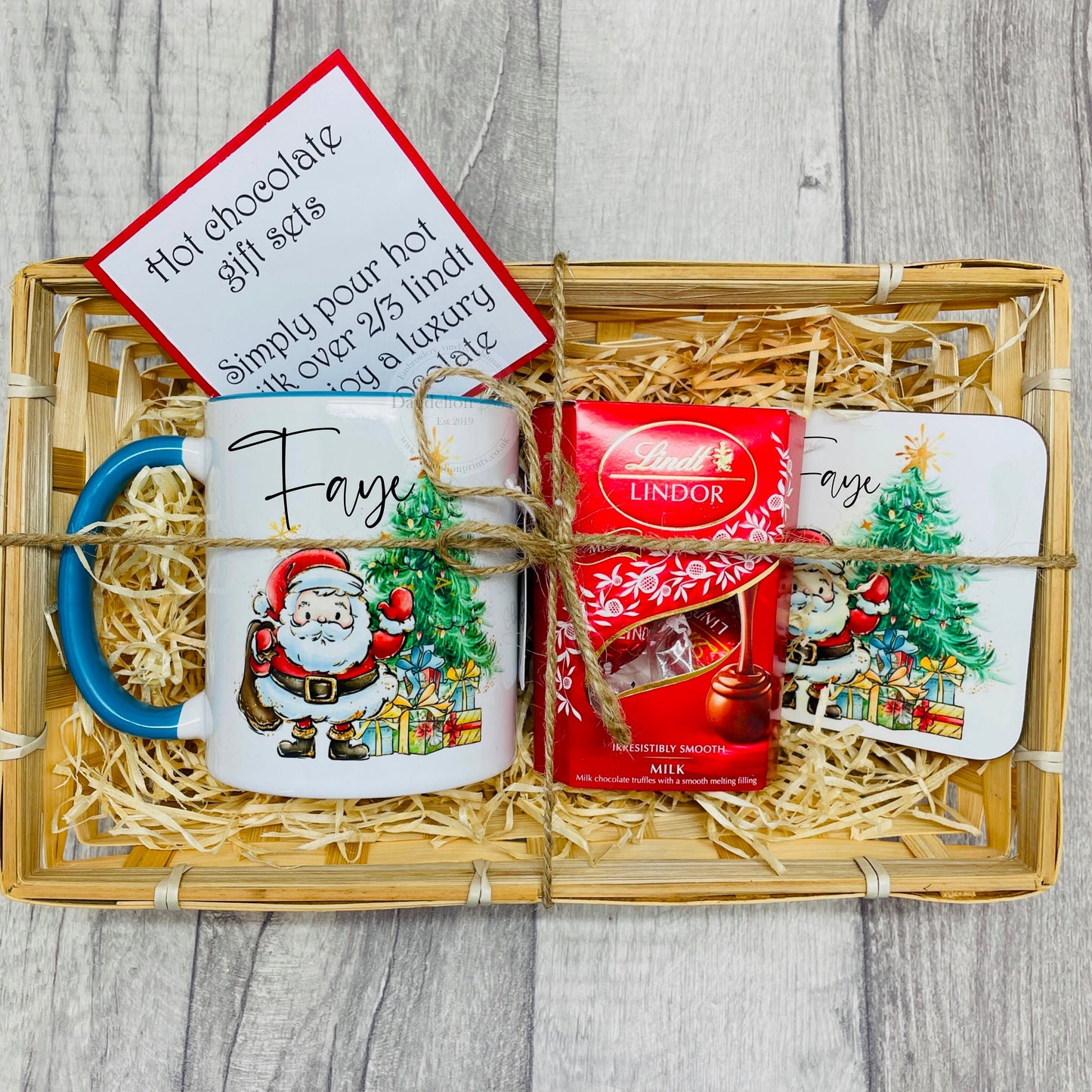 Christmas gift set in a wicker basket filled with a personalised mug, coaster and Lindt chocolates. The design on the mug features a cartoon Santa standing next to a Christmas tree with gifts below it. Ideal Christmas Eve gift. Stocking filler.