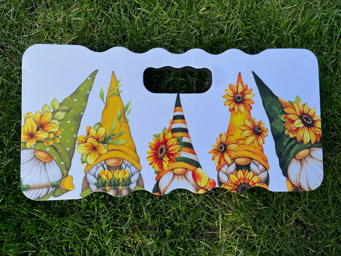 A soft kneeling pad laying on green grass photographed from above. The kneeling pad has a hand slot at the top with a colourful gnome design on the front. The design consists of 5 gnome / gonks all in autumn colours with sunflowers on their hats.