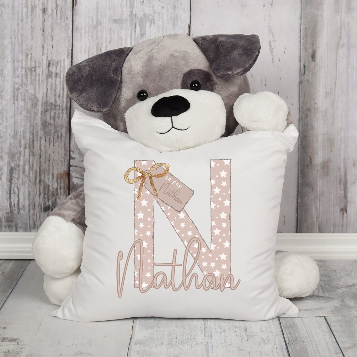 A pretty child cushion pictured in front of a large grey teddy dog both in front of a wood effect background. On the white cushion is a large initial N with the name Nathan below. The initial is filled with a starry design.
