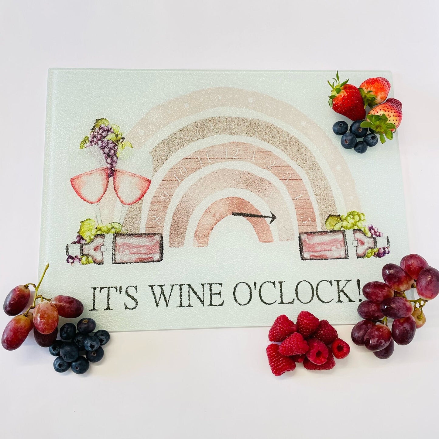 A glass chopping board pictured from above on a white background. Pictured with fruit for display. The design printed on the chopping board is a picture of a rustic dial with the needle to the right with words "its wine o'clock'