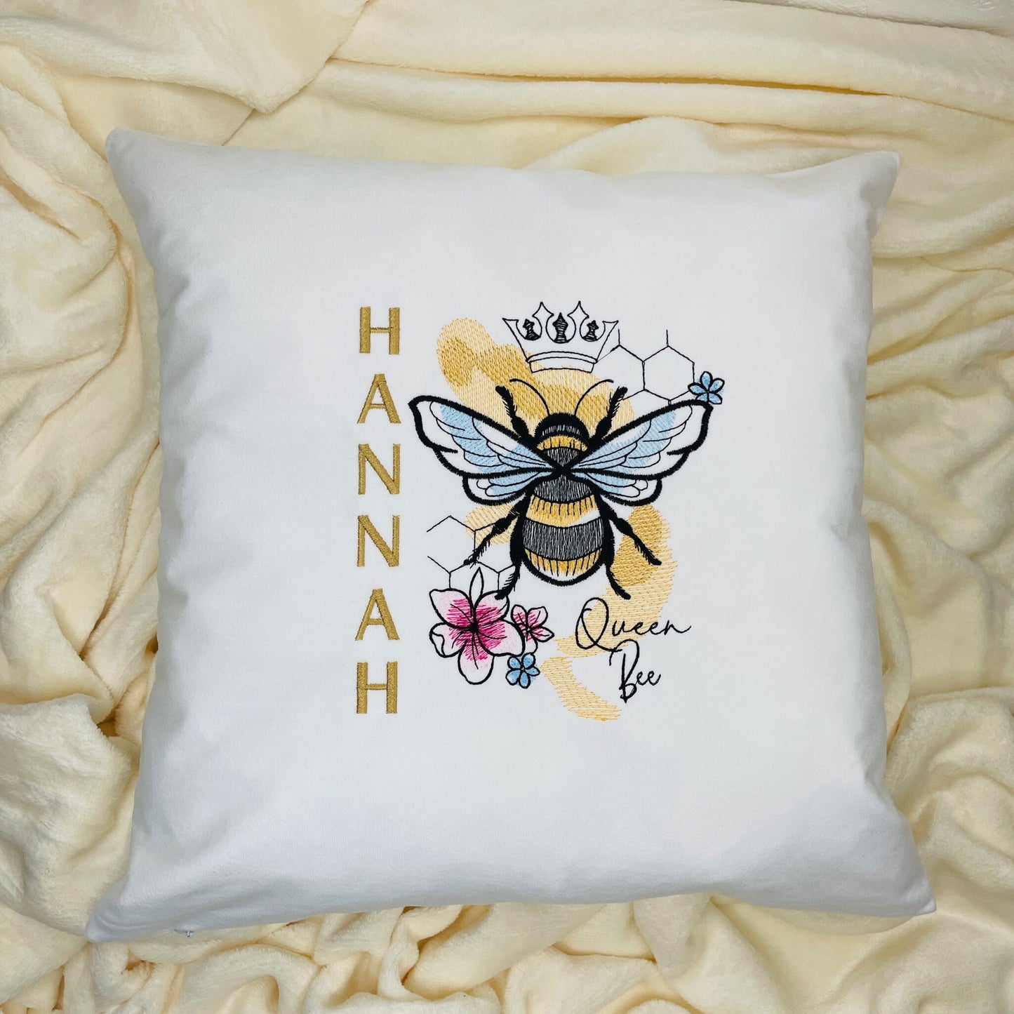 This is a white cushion with a beautiful bee embroidered onto it. The bee is decorated with stitched honeycomb, pretty florals and a crown. There is text to the right saying ‘Queen bee’ and a persons name downwards on the left.