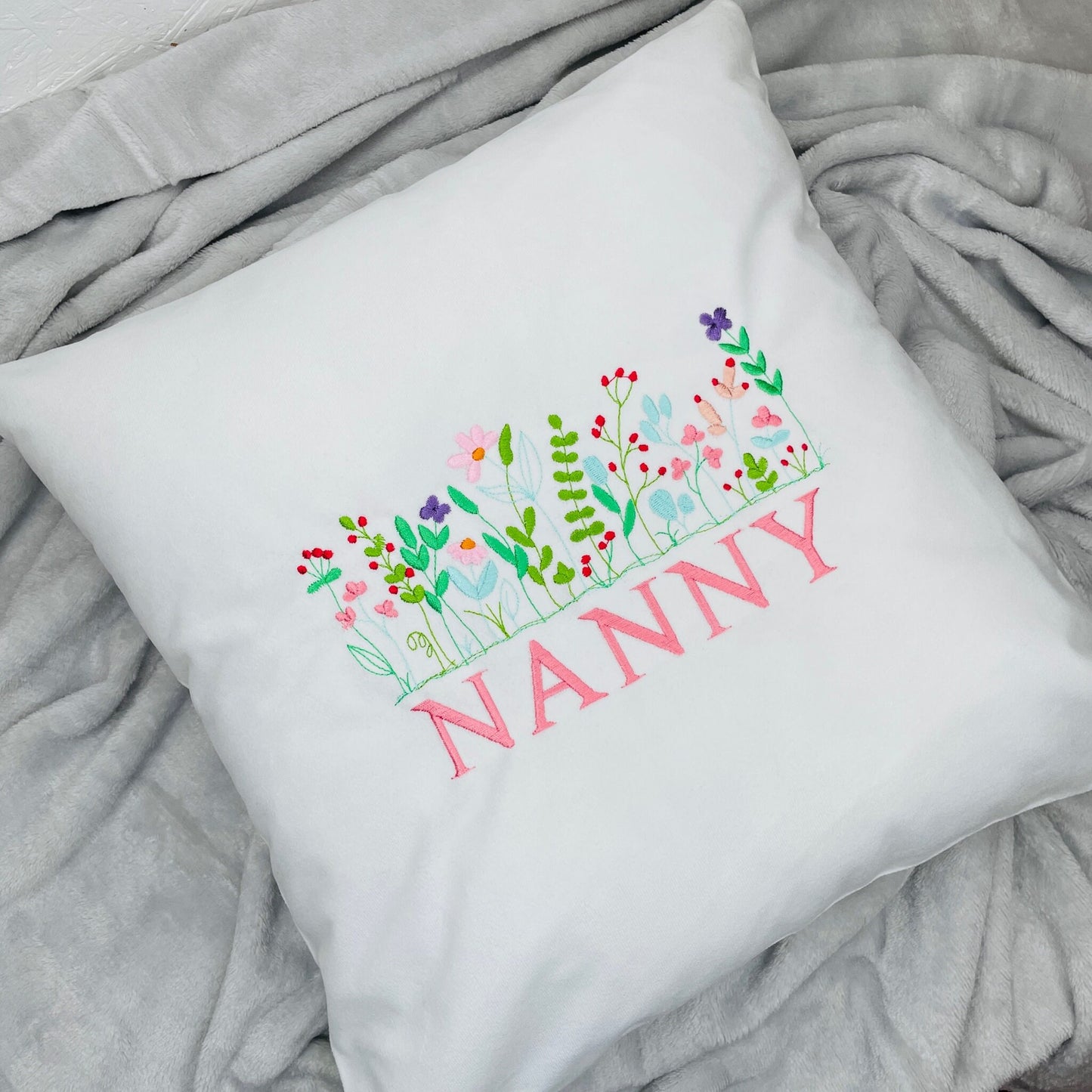 A beautiful but dainty wildflower design embroidered onto a super soft white velvet cushion. Flowers are pink, purple, red, cream and green leaves. Below the wildflowers is a name embroidered on.