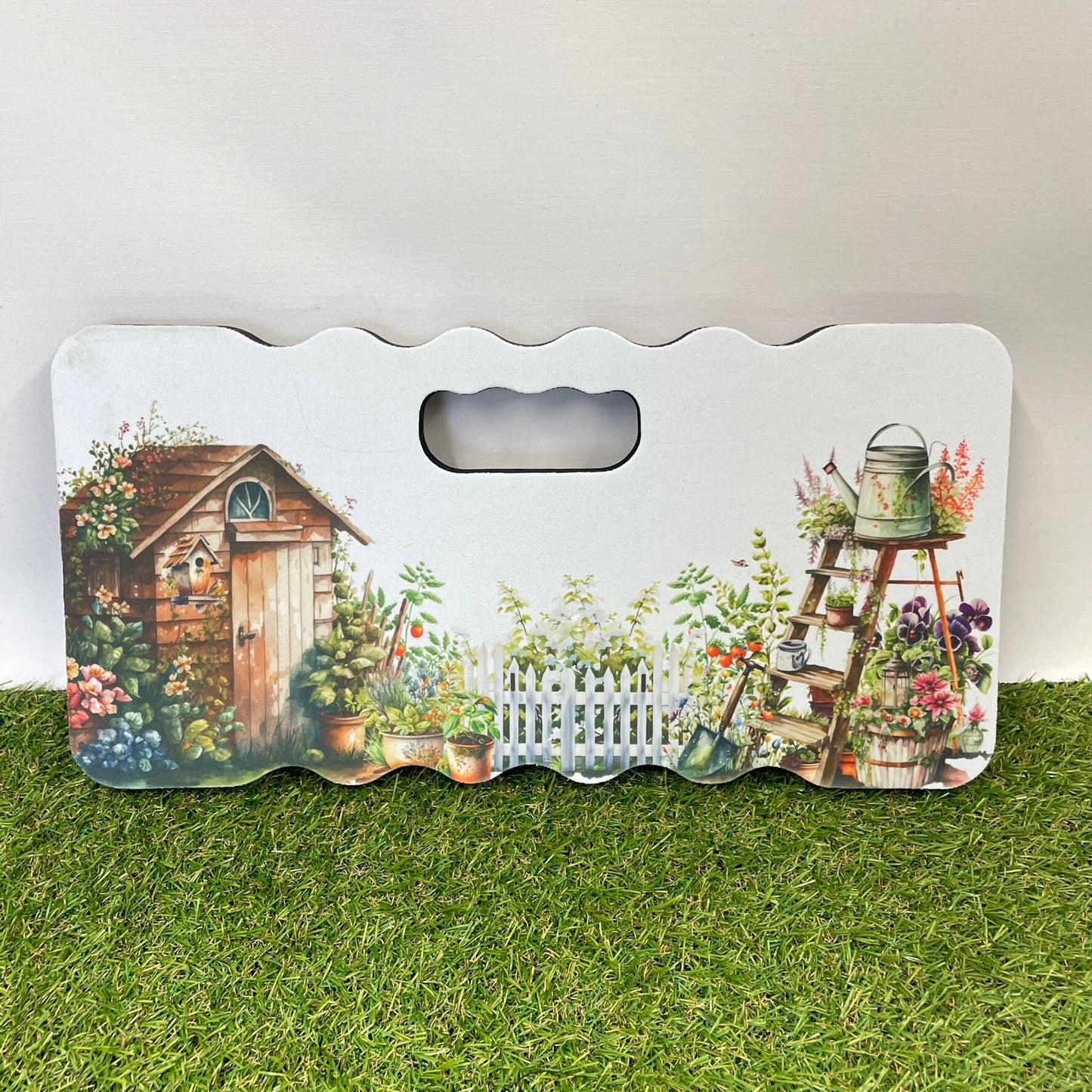 This is a garden kneeling pad printed with a beautiful busy garden. To the left is a wooden shed surrounded by plants. On the right is a set of wooden ladders with flowers trailing. Lots of plants along the middle personalised with a name at the top