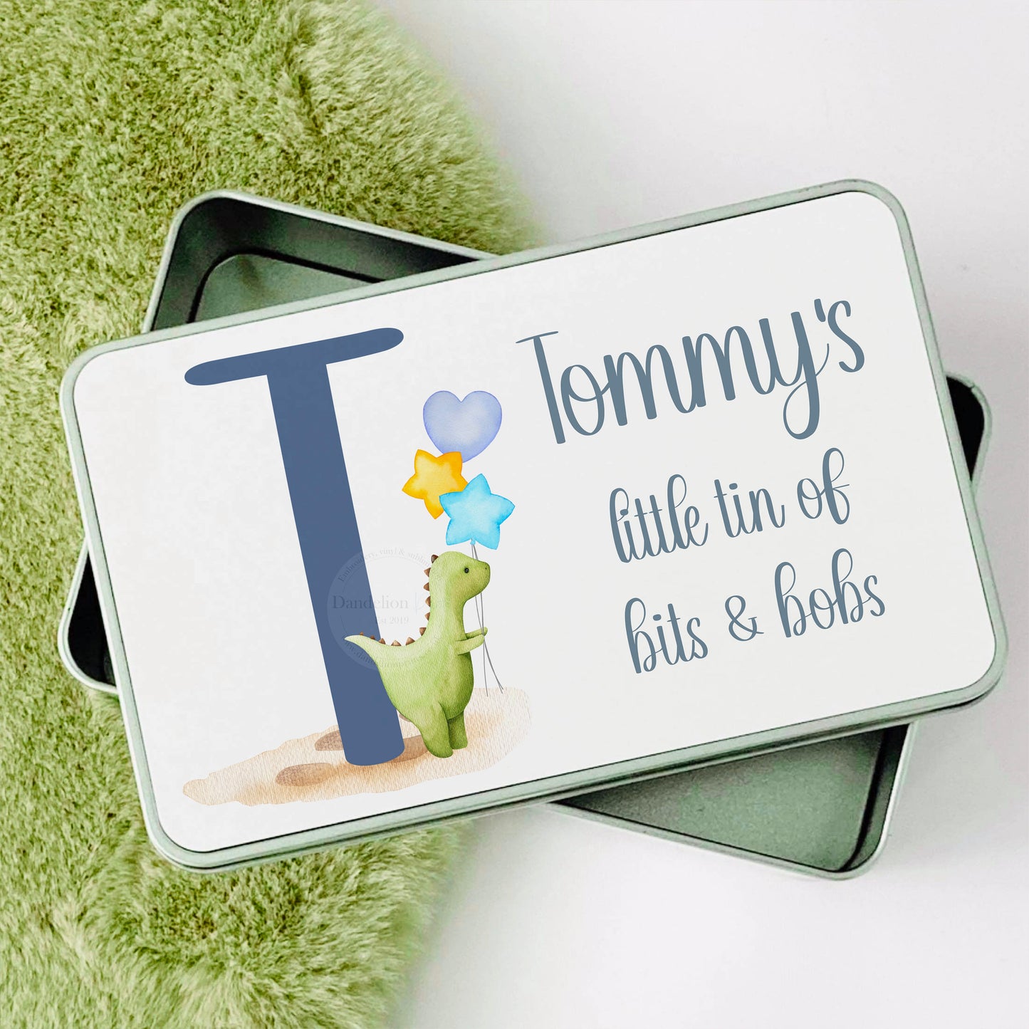 This is a medium sized metal storage and accessory tin. Printed on the top is a cute watercolour green dinosaur with balloons next to a blue initial. There is a name printed above the Dino. Personalised gift for boys and babies