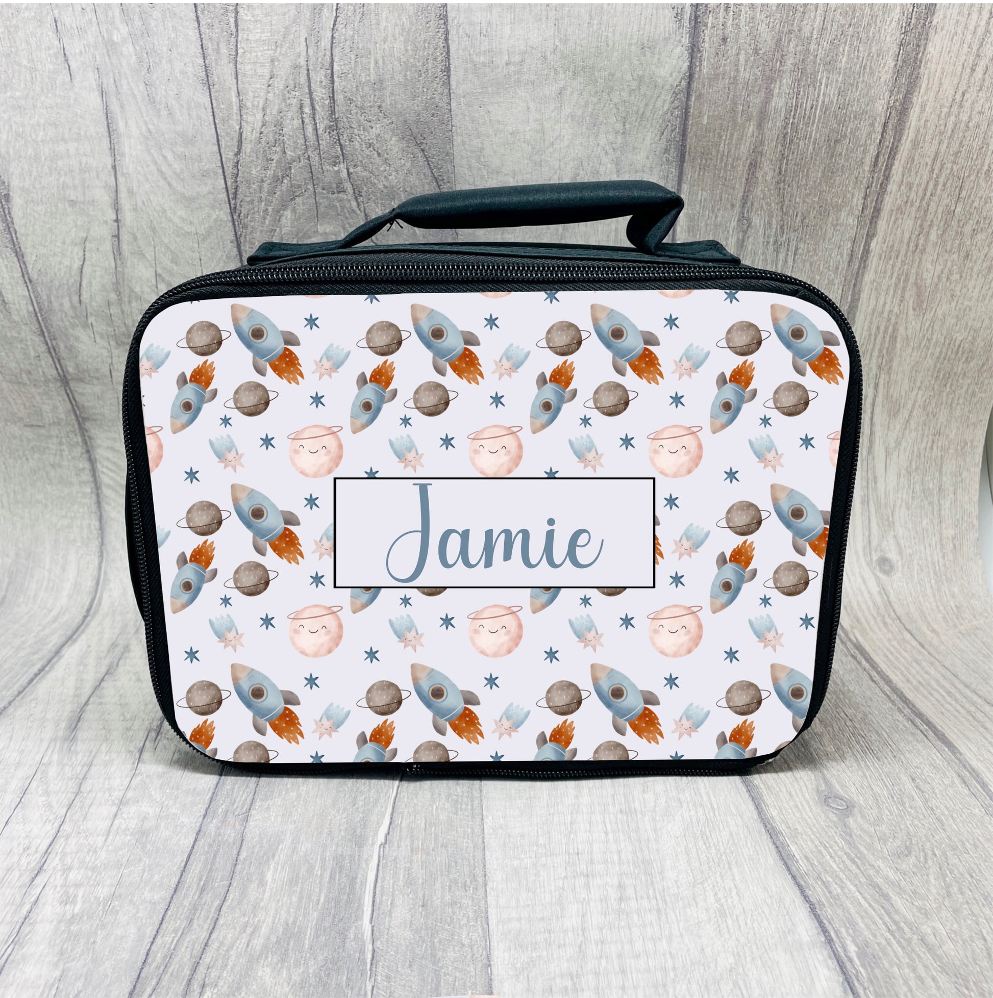 This is a black base lunch box with a handle at the top. The bag is fully insulated with a full zip closure. Printed with a pattern of rockets and space items and also a name in the middle.