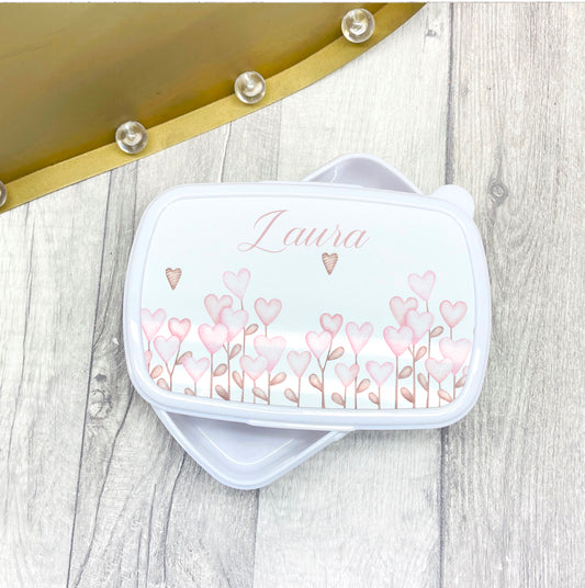 This is a white lunch box with separate lid. The lid is printed with a border of pretty pink heart flowers with a name above it. These are great for school or nursery.