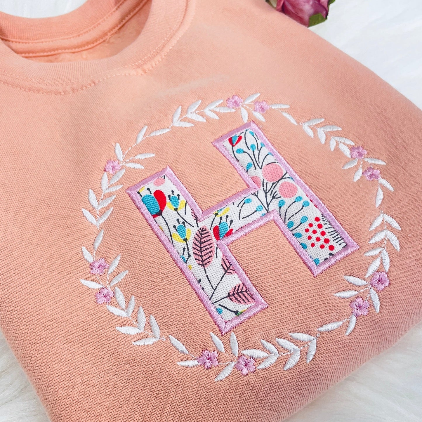 This is a pink sweater with a fleece lining. The sweater has lovely detailed pink and white embroidery on the chest featuring a floral wreath. Inside the wreath is a capital letter of you choice with a floral pattern inside and a thick embroidered outline.