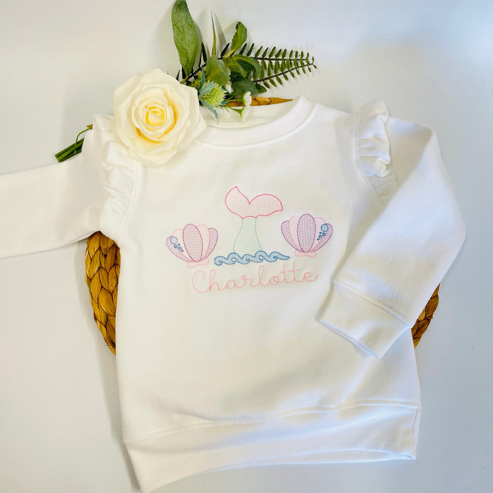 This is a white sweater with fleece lining. It is super soft but also fashionable with the frilled shoulders. The sweater is embroidered with a mermaid style sketch stick consisting of a mermaid tail between two sea shells in pink and purple theme. A name is embroidered below the design.