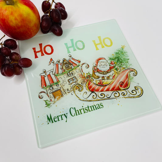 This is a square glass chopping board printed with a Christmas design. This design consists of a cartoon Santa on his sleigh. Santa is sitting on a pile of gifts in front of a festive house, giving the impression he is about to deliver the gifts.