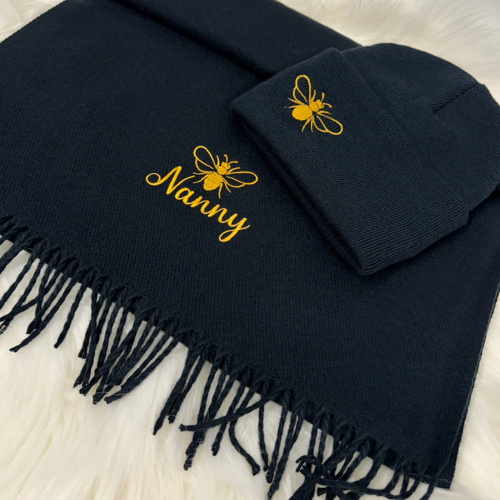 This is a navy hat and scarf set. Embroidered on the hat to the side is a golden bee design. On the scarf is also the matching bee design with the word nanny below also in gold