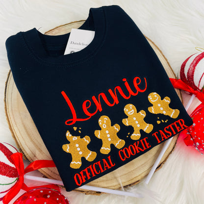 The embroidered design on the front of this sweater consists of four gingerbread men, two have been bitten but they all have a funny little face. The design has a name embroidered above it and 'Official cookie taster' embroidered below.