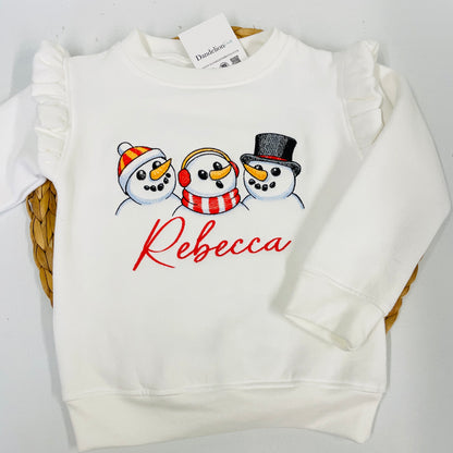 This is a beautiful white filly sleeve sweater for girls. Seasonal snowmen are featured embroidered on the front wearing different accessories. A name is embroidered below the images. Keep your little ones fashionably warm this winter with a stylish sweater