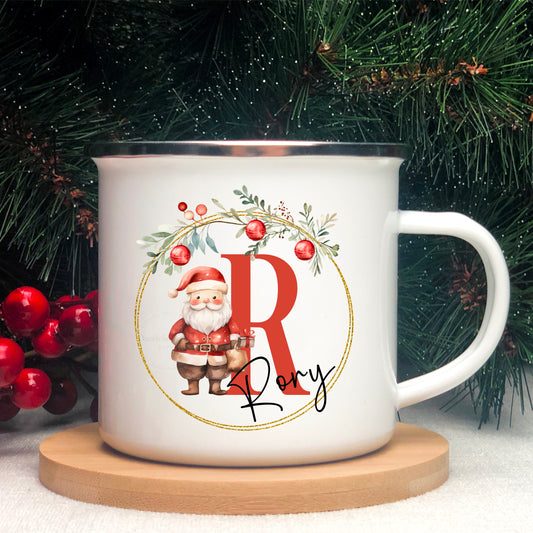 This is a white enamel mug with silver rim. To the front is the design. This consists of a Christmas garland with a Father Christmas figure to the centre. There is an alphabetical letter to the right with the full name across the bottom.