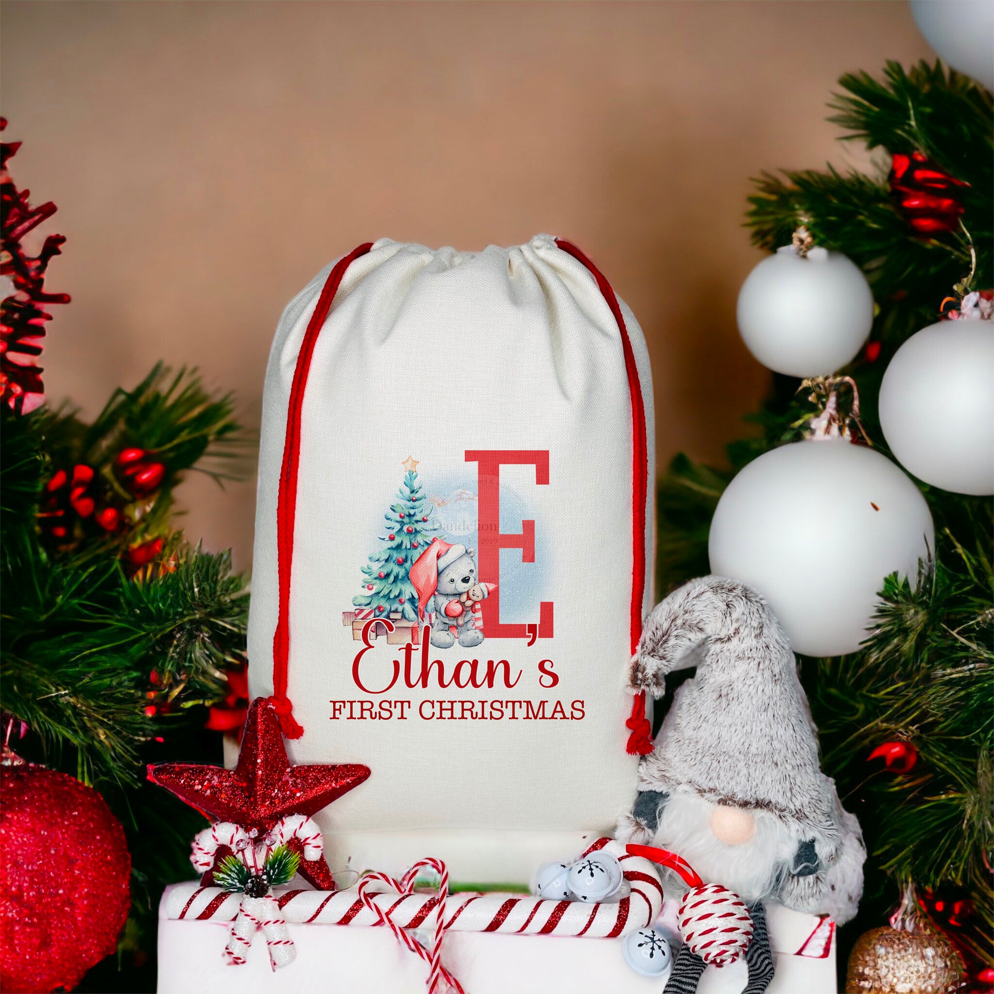 Here is a Christmas sack printed with a teddy bear wearing a Santa hat holding a toy in front of a blue Christmas tree with presents next to a large initial. There is a blue background and a moon. Below it says My First Christmas with a name.