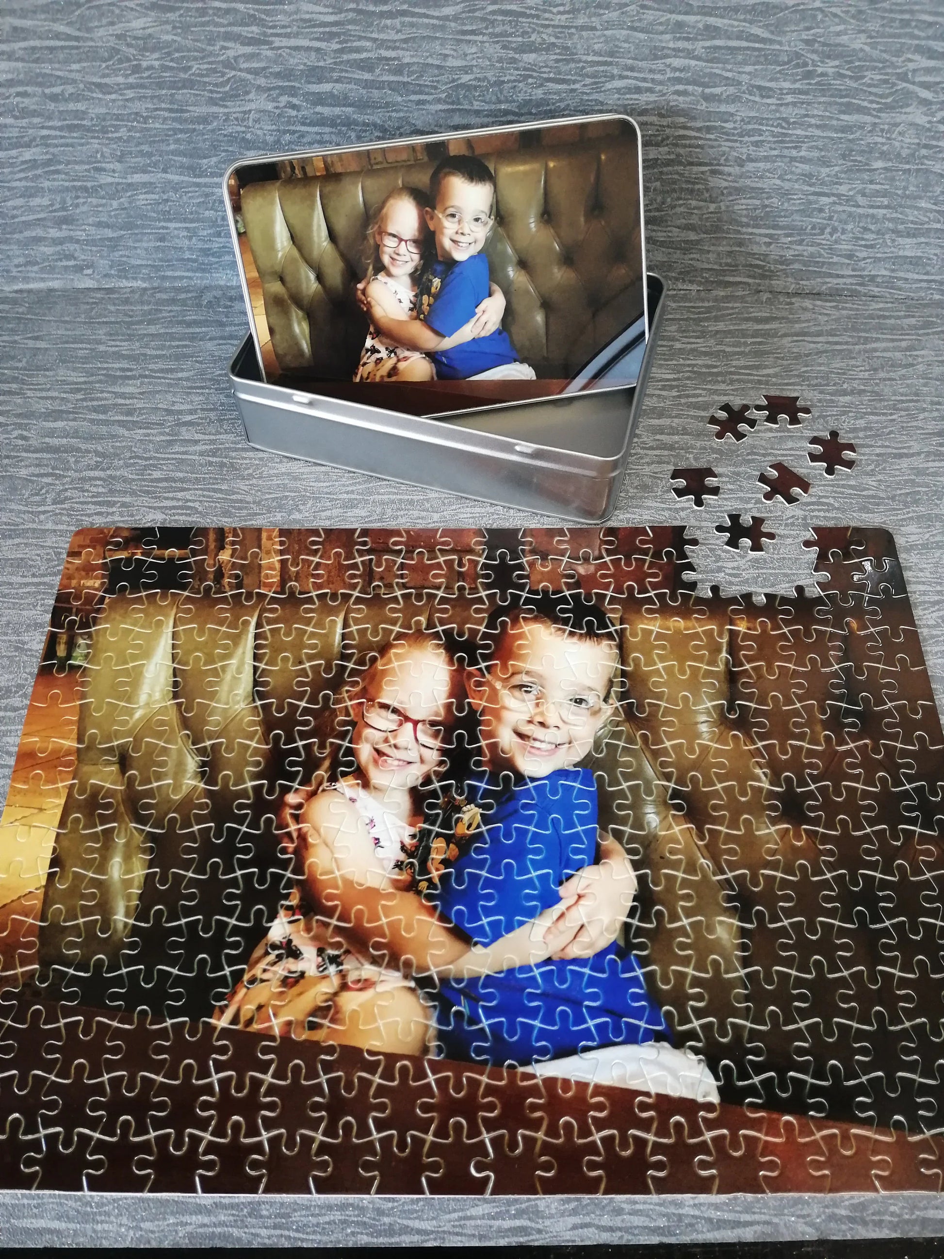 This is a photo puzzle with a printed storage tin. The puzzle has been printed with an image of two children. The print goes edge to edge. The lid of the tin is also printed with the same image. The puzzle is made with a single flower to the side.