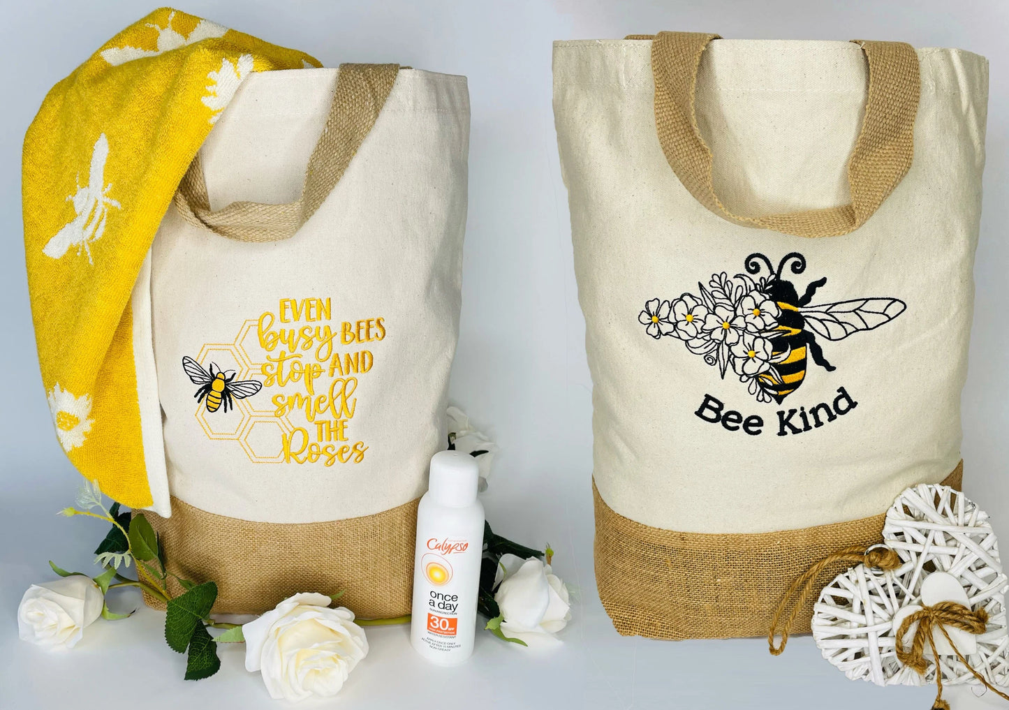 Image is of two cream tote bags. Each has an embroidered design. The base and handles are a light brown hessian material. One design is a large Bee with the words Bee Kind. The other a yellow bee poem. Photo with a yellow towel and sun cream.