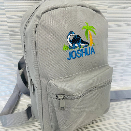 This is a grey backpack with a diplodocus, palm tree and grass embroidered on the front. The dinosaur is very detailed. There is a name embroidered below the dinosaur. These bags are also available in pink, navy, sky blue, sage, mint and lilac.