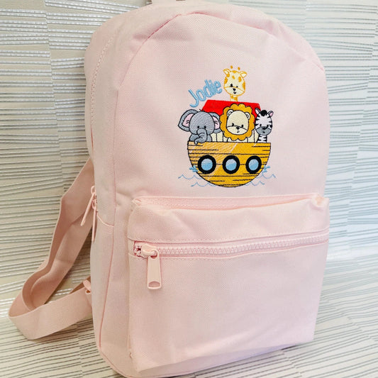 This is a pink backpack with an animal arc embroidered on the front. The arc is brown and red and is carrying a giraffe, elephant, lion and zebra. There is a name embroidered at the top of the design. Bags also available in other colours