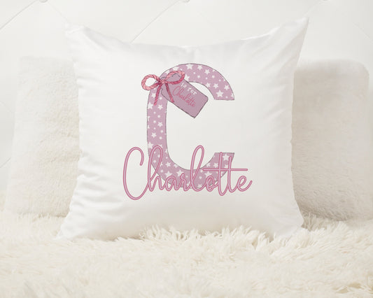 This is a 40x40cm white velvet book cushion. The cushion is printed with a pink initial full of stars. It also has a tag and bow on it. The design is finished with a name at the bottom.