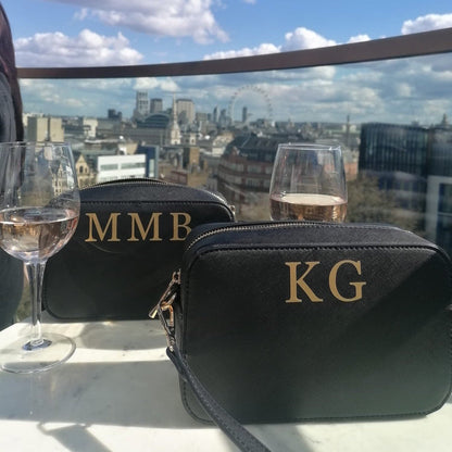 A black clutch bag photographed in front of an iconic London skyline. The bag is rectangle in shape with a matching black shoulder strip. There are initials KG on the top of the bag in gold vinyl.