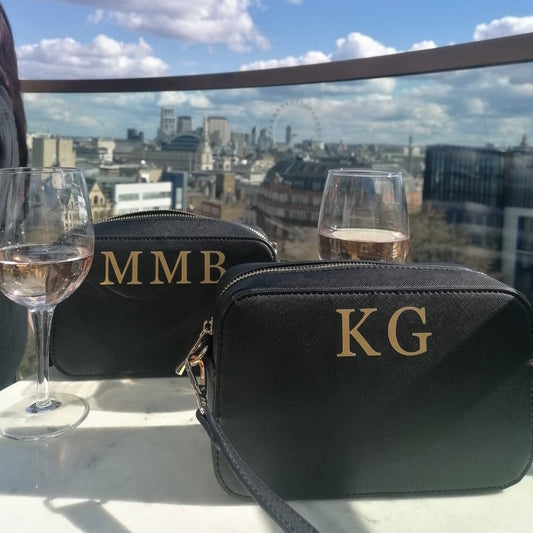 A black clutch bag photographed in front of an iconic London skyline. The bag is rectangle in shape with a matching black shoulder strip. There are initials KG on the top of the bag in gold vinyl.