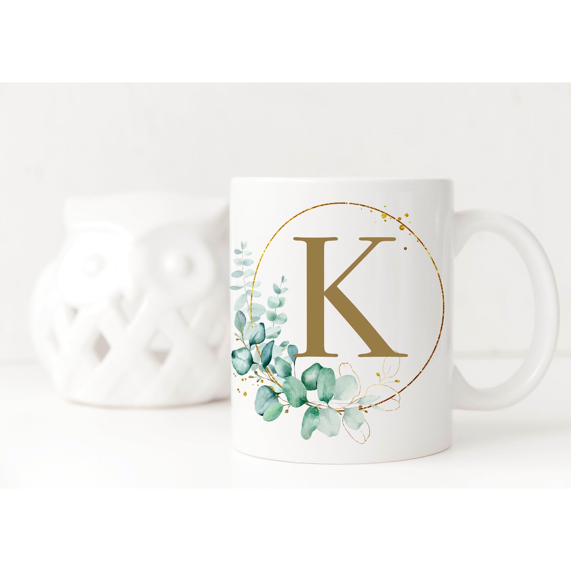 White mug displayed with an elegant print on it. The print is a eucalyptus hoop with pretty gold splashes and plenty of leaves. Inside the hoop is a gold initial.