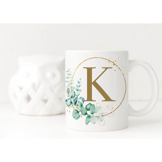 White mug displayed with an elegant print on it. The print is a eucalyptus hoop with pretty gold splashes and plenty of leaves. Inside the hoop is a gold initial.