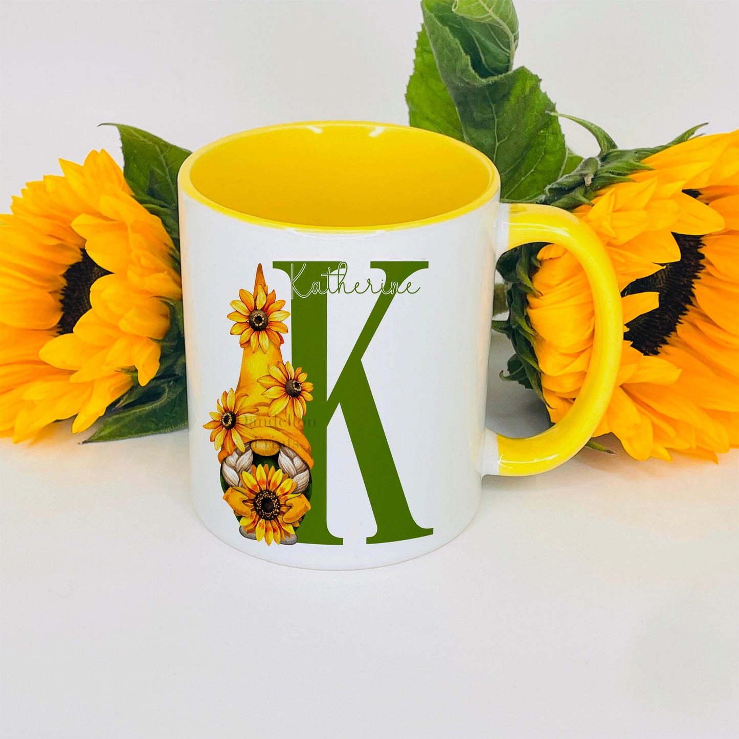 This is a white mug with a yellow handle and  yellow inside. The mug has an initial printed in green and a sunflower gonk standing next to it. The design also has a name printed across the top of the initial.