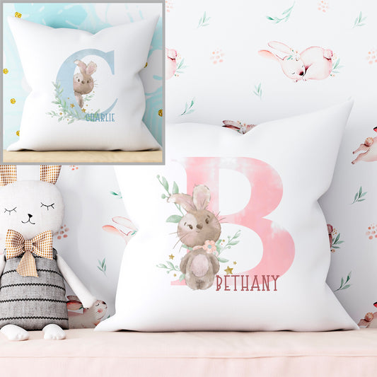 Super soft white cushion printed with a pink or blue bunny alphabet. The pink version has a pink cloudy initial and brown bunny wearing a pink bow. The blue version has a blue cloudy initial with a brown bunny. Both bunnies have pretty florals.